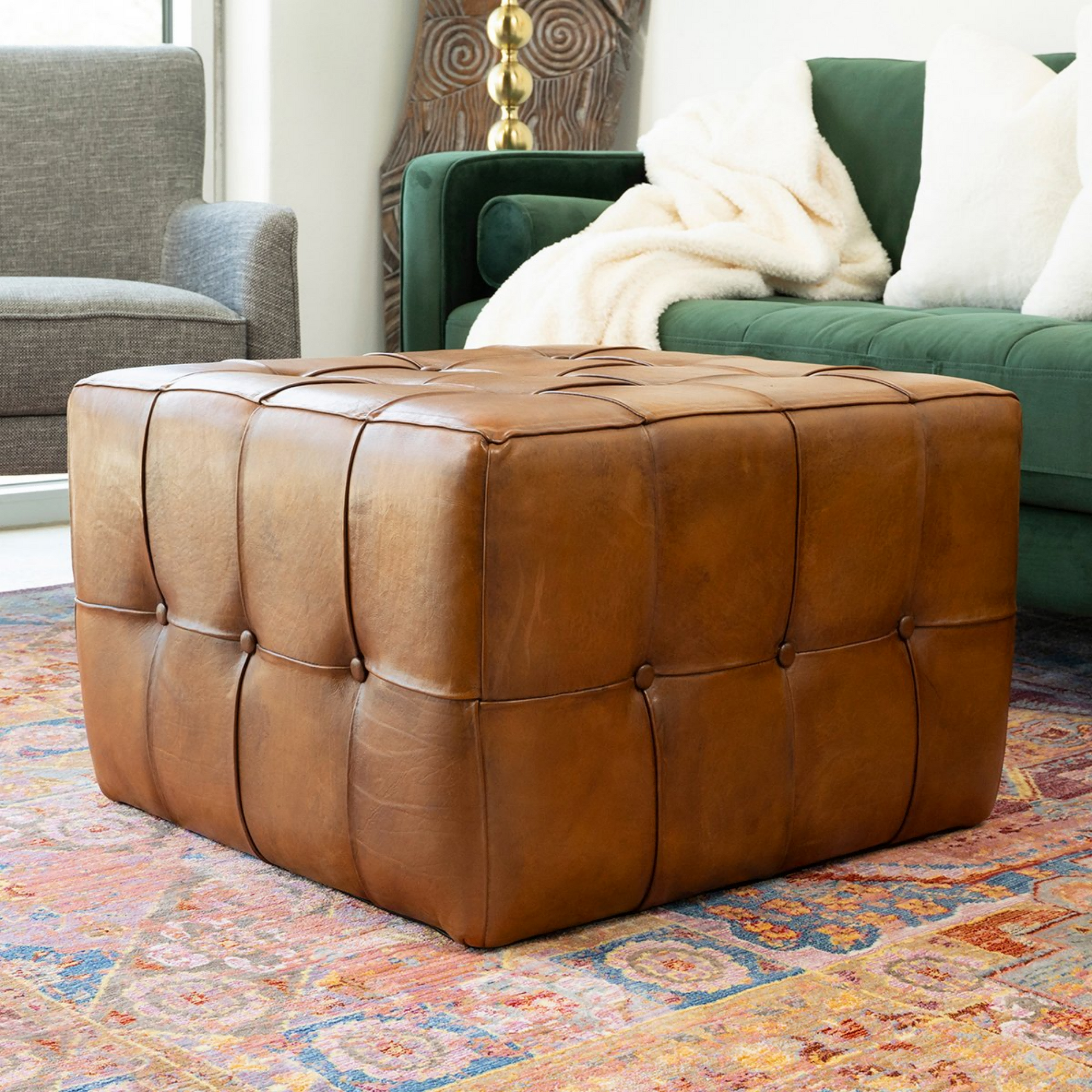 Tufted brown leather ottoman on patterned rug in living room with green velvet sofa