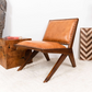 Modern brown leather accent chair with wooden frame on white rug, next to wood side table.