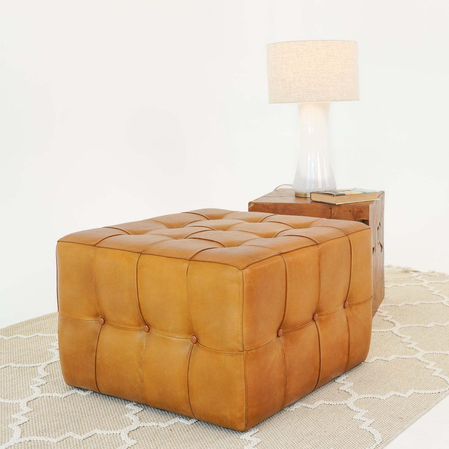 Tufted tan leather ottoman on patterned rug with wooden side table and white lamp, FredCo International furniture.