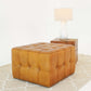Tufted tan leather ottoman on patterned rug with wooden side table and white lamp, FredCo International furniture.