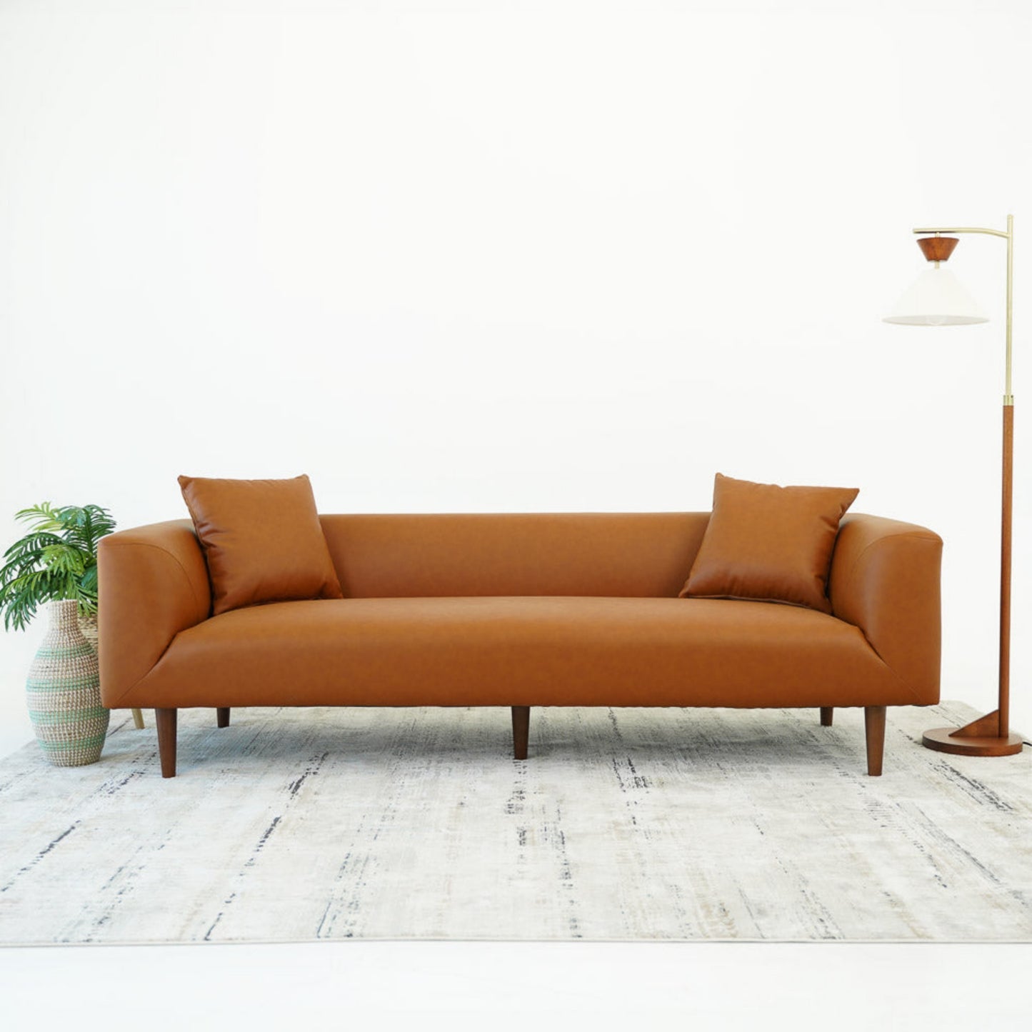 Mid-century modern brown sofa with two pillows on light rug, near potted plant and floor lamp