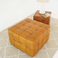 Tan leather tufted ottoman on patterned rug next to wood side table with lamp and book
