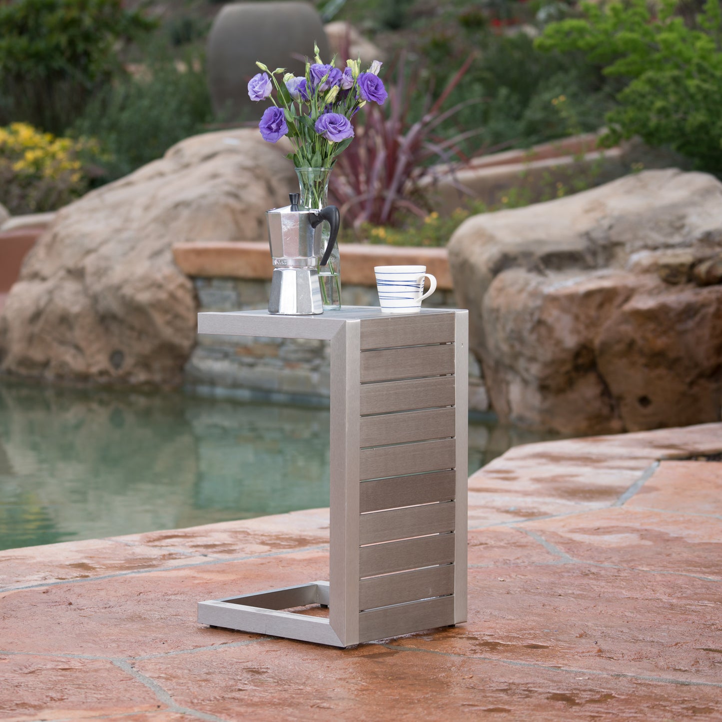 Modern outdoor side table with flowers, coffee maker, and mug by pool on stone patio