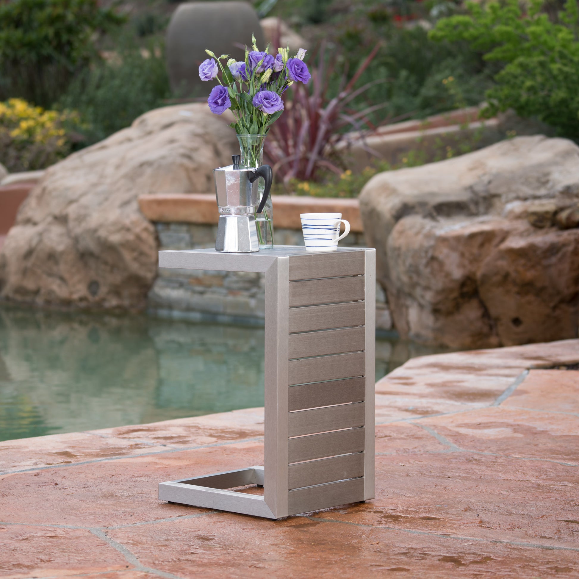 Modern outdoor side table with flowers, coffee maker, and mug by pool on stone patio