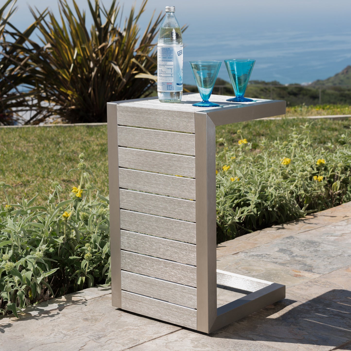 Modern outdoor side table with metal finish, blue glasses and water bottle, garden patio view
