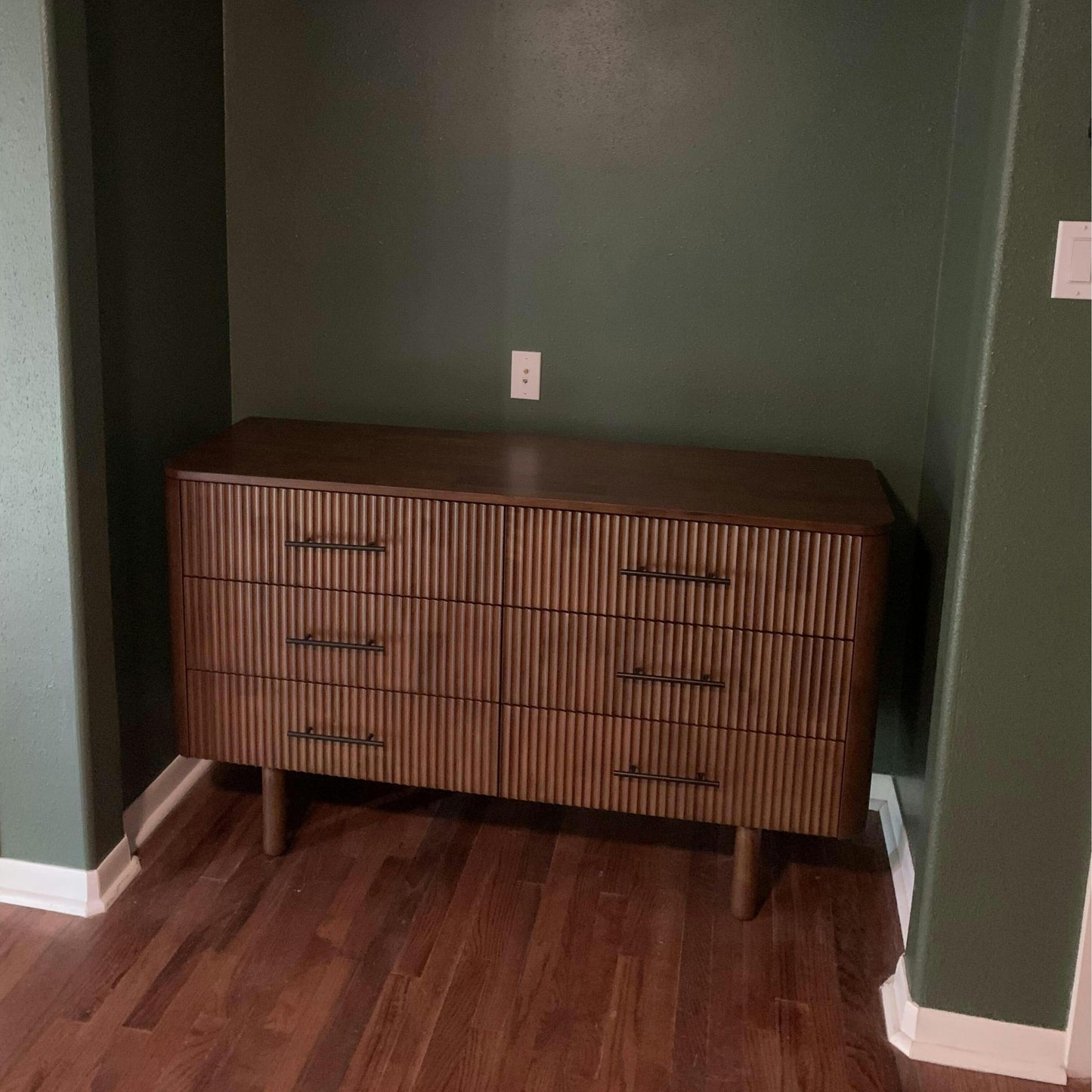 Mid-century modern wood dresser with vertical grooves, set against a green wall on hardwood floor.