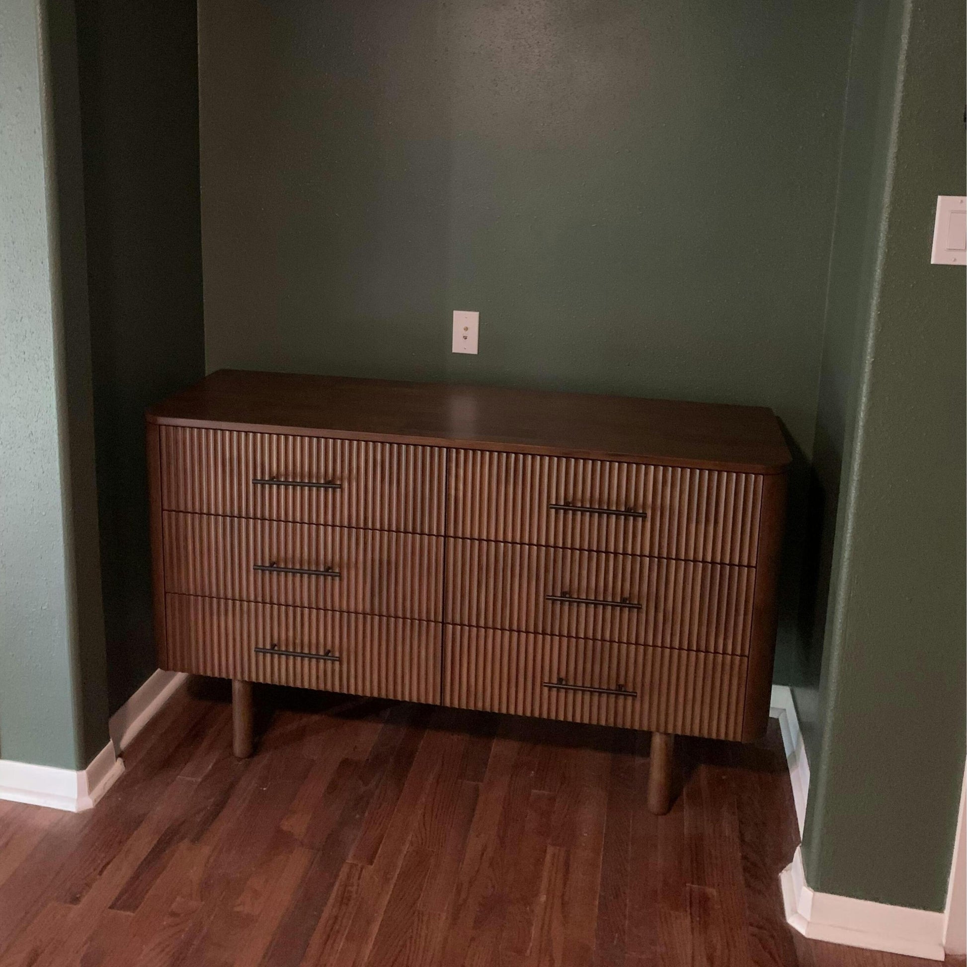 Mid-century modern wood dresser with vertical grooves, set against a green wall on hardwood floor.