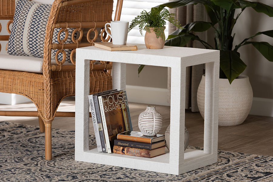 Modern white side table with books, vase, and plant, next to a rattan chair in a cozy living room.